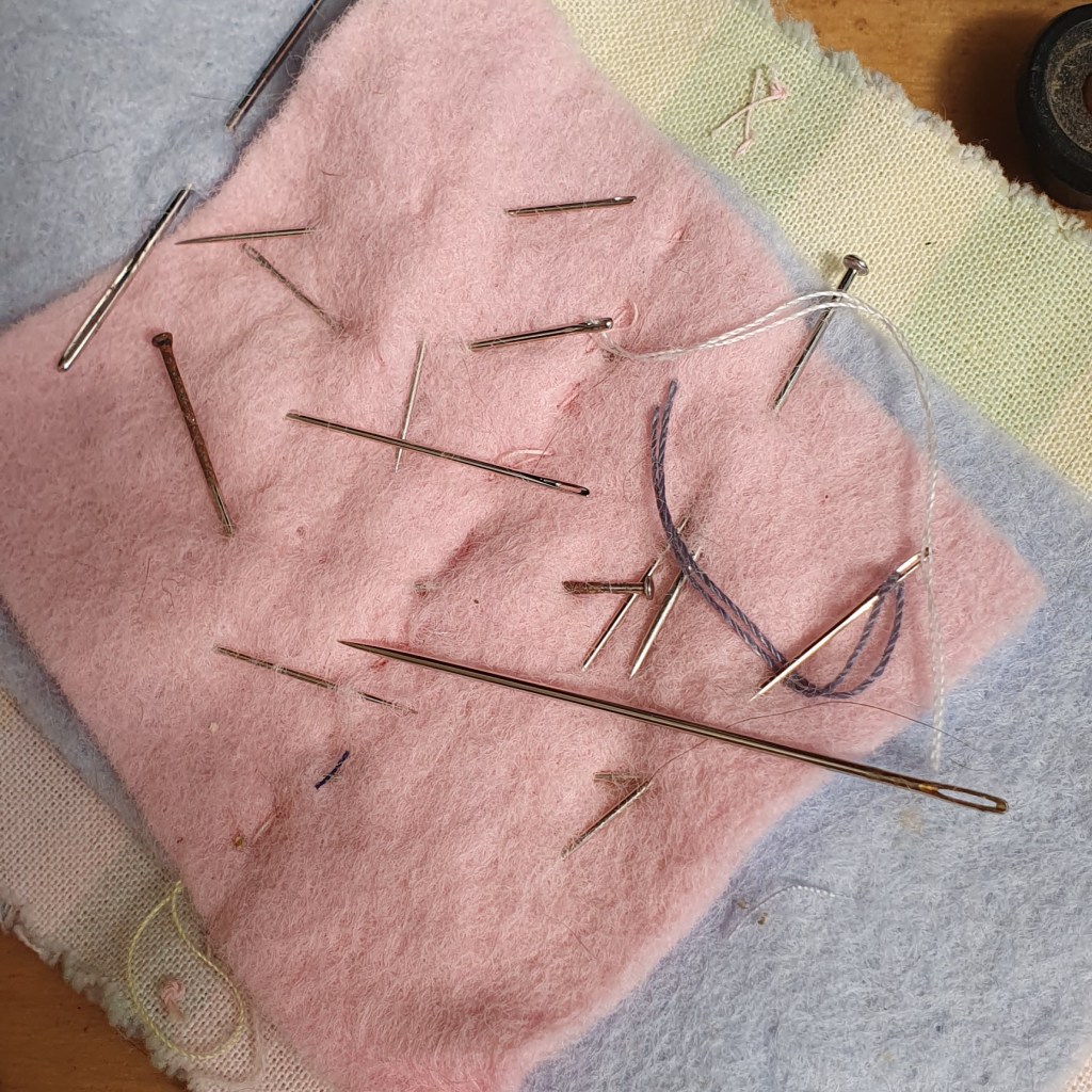 Various sewing needles arranged on a pink and blue felt surface.
